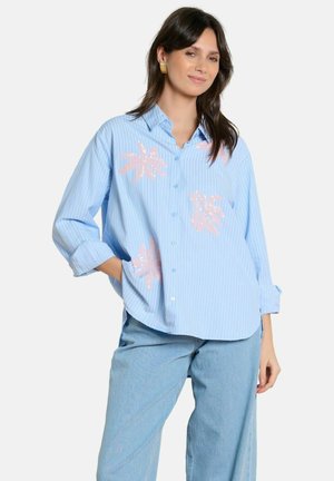 Frau trägt ein hellblau gestreiftes Hemd mit pinken Blumenmustern und hellblaue Jeans, posiert mit einer Hand in der Tasche.