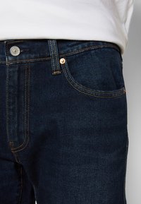 Jeans de mezclilla azul oscuro con costuras naranjas y botón plateado, usados con una camisa blanca metida por dentro, mostrando detalles del bolsillo delantero y los lazos del cinturón.