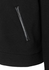 Schwarze Jacke aus strukturiertem Stoff mit einer eleganten, matt-schwarzen Reißverschlusstasche und einem gerippten Saum. Das Material weist ein dezentes Muster auf.