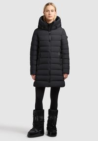 Black padded coat with a high collar and hood, featuring a zip front, side pockets, and a knee-length design. Paired with black snow boots.
