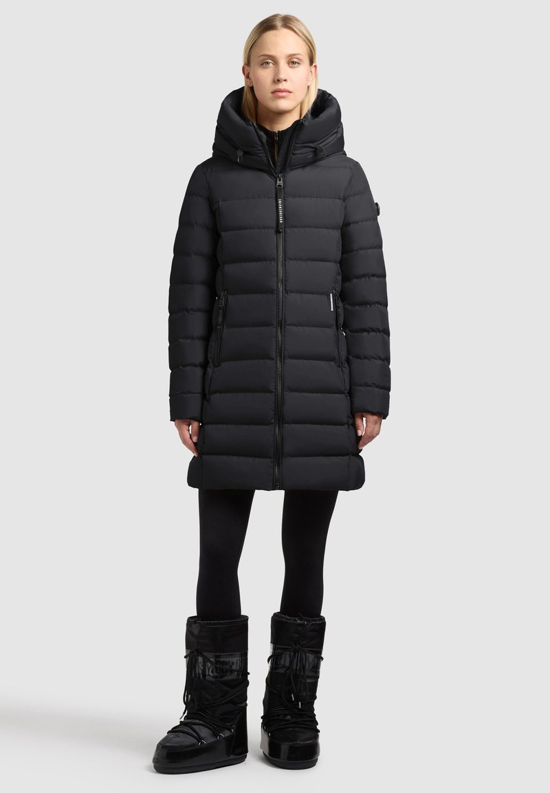 Black padded coat with a high collar and hood, featuring a zip front, side pockets, and a knee-length design. Paired with black snow boots.