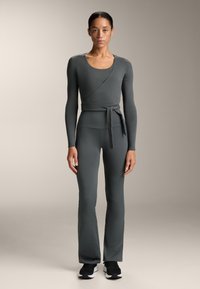 Siva dugmadna majica s dugim rukavima s vezanjem u struku i odgovarajuće flared tajice, napravljene od glatkog, rastezljivog materijala. Crne sportske cipele upotpunjuju izgled.