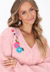 Rozā adīta džemperis ar ziedu izšuvumiem, rozā pogām un V veida kakla dizainu. Mīksta tekstūra, sarežģītas ziedu detaļas dažādās krāsās.