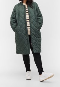 Gequilted groene lange jas met een ronde hals en zijzakken. Draagt over een gestreept sweater en zwarte broek, gecombineerd met witte sneakers.