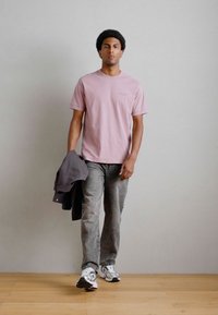 Ροζ κοντομάνικο μπλουζάκι, γκρι τζιν, γκρι μπουφάν ριγμένο στον βραχίονά του, λευκά αθλητικά παπούτσια με μαύρες λεπτομέρειες, στέκεται μπροστά σε ουδέτερο φόντο.