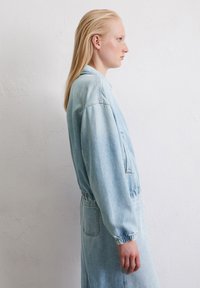 Person mit langen blonden Haaren, der nach rechts schaut, trägt eine hellblaue Denimjacke vor einem schlichten weißen Hintergrund.