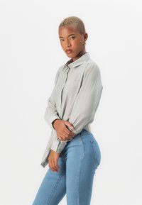 Long-sleeve button-up shirt in light grey with thin white stripes, featuring a collar and chest pocket, paired with fitted light blue jeans.