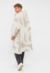 Beige regnjacka med huva, stora abstrakta ljusgrå mönster, överdimensionerad silhuett, elastiska ärmslut och sidofickor, matchad med mörka jeans.