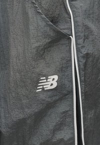 Υφαντό από σκούρο γκρι ύφασμα με λευκή περιμετρική ραφή και λευκό λογότυπο New Balance σε κοντινή λήψη αθλητικού μπουφάν.