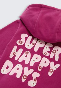 Haut à capuche fuchsia présentant de grandes lettres roses avec l'inscription "JOURS SUPER HEUREUX", rehaussé de détails floraux blancs et pailletés. Texture douce.