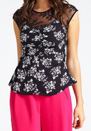 Femme portant un haut en dentelle florale noire transparente avec un motif floral blanc et un pantalon rose vif, visible du cou aux hanches.