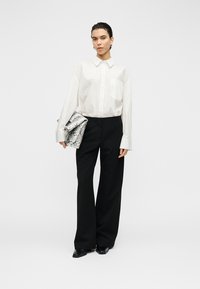 Chemise blanche à boutons avec une poche, pantalon noir à jambes larges, et une pochette à motif de peau de serpent. Des chaussures noires complètent la tenue.