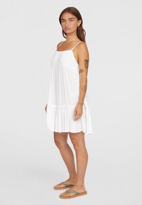 Weißes, ärmelloses Kleid mit plissierter Textur, einem taillierten Oberteil und einem ausgestellten Rock, der mit einem rüschierten Saum abgeschlossen ist. Sandalen mit einem grünen Riemen.