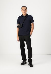 Námořnická polokošile s bílými akcenty, černá přes shoulder taška, černé kalhoty a černé boty. Model stojí na neutrálním pozadí.
