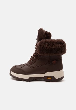 Bota de invierno de cuero marrón oscuro con forro grueso de piel, cordones en la parte delantera y suela Vibram blanca sobre un fondo claro.