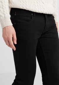 Black denim jeans with a slim fit, visible pocket details, and contrasting stitching. Fabric appears soft with a slight sheen.