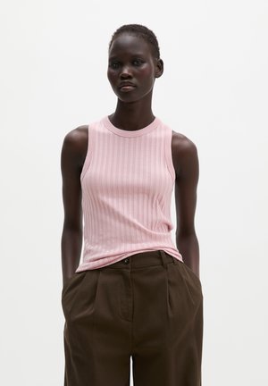 Donkere vrouw die een mouwloze roze geribde top en een hoge taillebruine geplooide broek draagt, met handen in de zakken.
