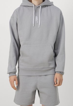 Personne portant un hoodie gris clair avec poche avant et cordons, associé à un short gris assorti, se tenant devant un fond uni.