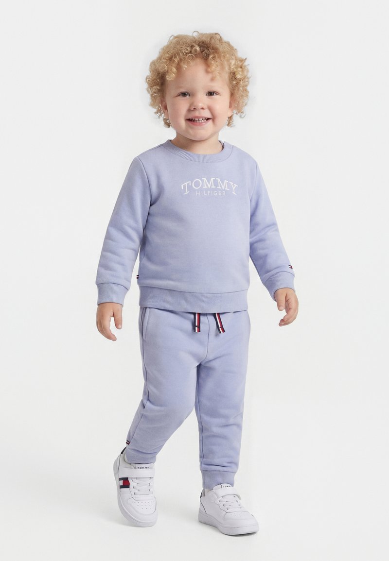 Χαμογελαστό νήπιο με ξανθά σγουρά μαλλιά που περπατά, φορώντας ανοιχτό μωβ φούτερ Tommy Hilfiger και ταιριαστά παντελόνια φόρμας, λευκά αθλητικά παπούτσια με λογότυπο.