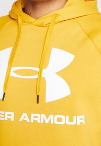 Sudadera amarilla con bolsillo canguro, cordones y un logo blanco de Under Armour en el frente. Tela suave y texturizada con un ajuste relajado.
