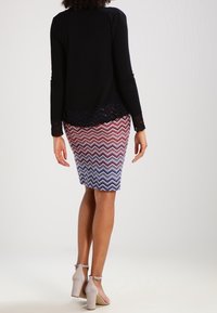 Černý top s dlouhým rukávem a krajkovým lemem, doplněný tužkovou sukní s vícenásobným cikcak vzorem v červené, bílé a modré; zvýšené béžové podpatky.