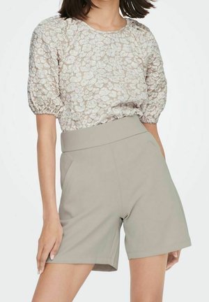Femme portant un short beige taille haute avec poches et un chemisier fleuri à manches courtes avec manches bouffantes.