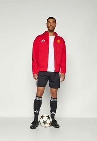 Red zip-up jacket with Manchester United logo, white t-shirt, black shorts, black socks with white stripes, and black soccer shoes, standing by a soccer ball.