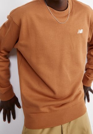 Sweat-shirt marron à col rond et poignets côtelés, avec un petit logo blanc sur le côté gauche de la poitrine. Texture de tissu lisse.