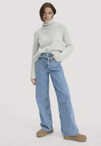 Eine Frau trägt einen hellgrauen, gerippten Rollkragenpullover, hochgeschnittene, lockere blaue Jeans mit einem gebundenen Gürtel und beigen Plateauschuhen und steht entspannt.
