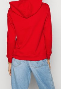 Sudadera con capucha roja que presenta una textura suave, mangas largas y puños acanalados, combinada con vaqueros de mezclilla azul claro.