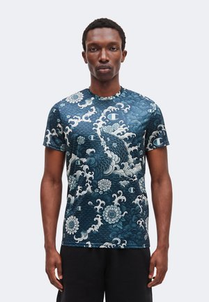 Man met een donkerblauw shirt met korte mouwen met witte bloem- en golfpatronen, staand tegen een effen lichte achtergrond.