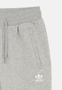 Pelēki sporta bikses, kas izgatavotas no mīksta auduma, ar elastīgu jostu, divām sānu kabatām un baltu Adidas logotipu apakšējā kreisajā malā.