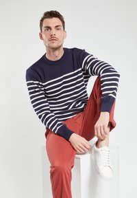 Pull bleu marine avec des rayures horizontales blanches, poignets et col côtelés, associé à un chino rouge et des baskets blanches, assis sur un bloc blanc.
