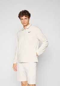 Light beige zip-up jacket with a collared neck and black Nike logo on the left chest, paired with matching beige shorts. Smooth texture.