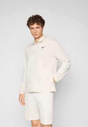 Lys beige zip-up jakke med krage og svart Nike-logo på venstre bryst, sammen med matchende beige shorts. Glatt tekstur.