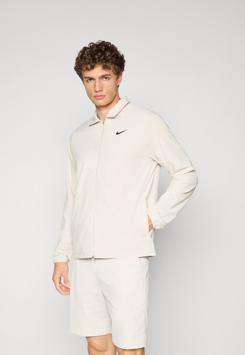 Light beige zip-up jacket with a collared neck and black Nike logo on the left chest, paired with matching beige shorts. Smooth texture.