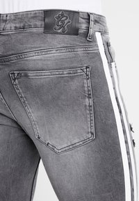 Jeans de denim gris con un parche de cintura de cuero negro, franjas laterales blancas y un detalle desgastado en el bolsillo trasero.