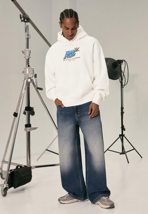 Hvid hættetrøje med blå og rød logo, parret med brede, falmede blå jeans. Outfitet komplementeres af grå sneakers. Neutral baggrund.
