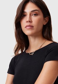 Gullkjede med to lagdelte kjeder og et geometrisk anheng. Brukes over en svart kortermet topp, glatt tekstur og minimalistisk design.