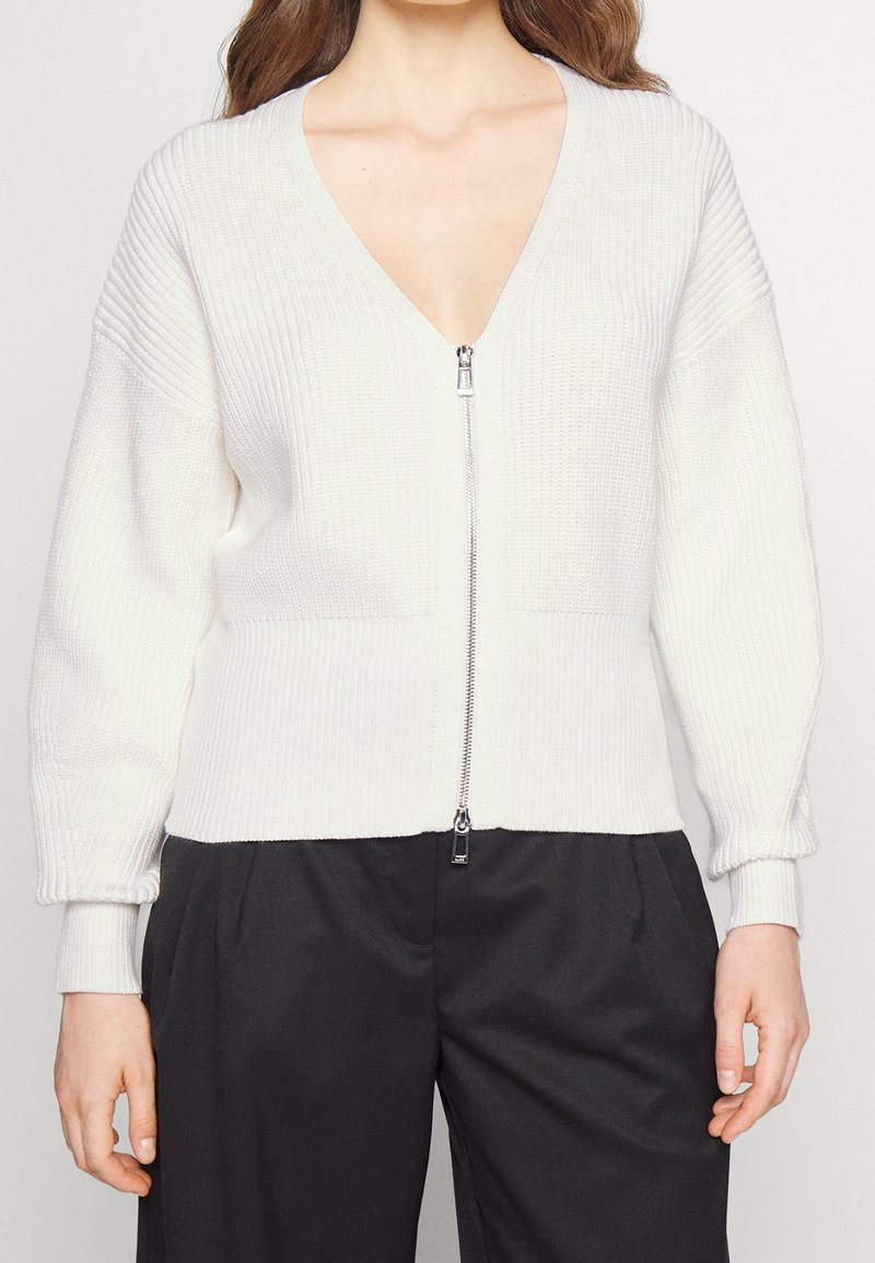 Femme portant un cardigan zippé blanc côtelé à manches longues et un pantalon noir taille haute, vue du cou jusqu'aux hanches.