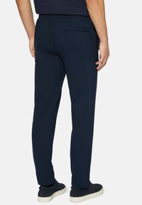 Man som bär marinblå casualbyxor och svarta låga sneakers står mot en enhetlig vit bakgrund.