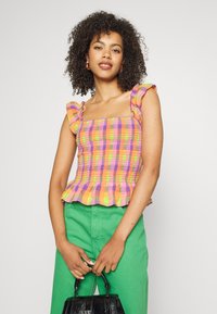 Top colorido com pregas e mangas com babados, apresentando um padrão quadriculado em laranja, rosa e amarelo, combinado com calças verdes e uma bolsa preta.