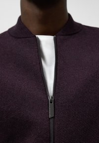 Ciemnofioletowa bombka dzianinowa z zamkiem błyskawicznym z przodu i okrągłym kołnierzem, noszona na białej koszulce. Teksturowany materiał i metalowy zamek.