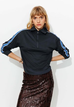 Femme blonde portant un sweat à demi-fermeture éclair noir avec des rayures bleues sur les manches et une jupe brune foncée à sequins, posant les mains sur les hanches.