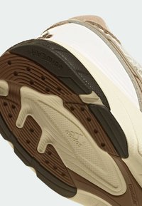 Σόλα παπουτσιού με υφή σε καφέ καουτσούκ, με αυλακώσεις και κυκλικά σχέδια. Άσπρη μεσαία σόλα με λεπτομέρειες λογότυπου και μπεζ τόνους κοντά στη φτέρνα.