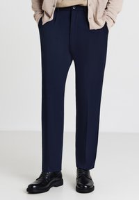 Pantaloni blu navy con un design a gamba dritta, texture liscia, due tasche frontali e chiusura a bottone. Indossati con scarpe eleganti nere.