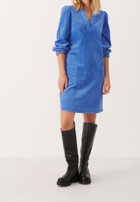 Abito in corduroy blu con scollatura a V e maniche a palloncino, abbinato a stivali di gomma neri al ginocchio. L'abito presenta un tessuto strutturato.