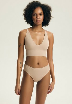 Kobieta o kręconych włosach, ubrana w beżowy prążkowany biustonosz i majtki, stoi z neutralnym wyrazem twarzy na jasnym tle, pokazując tatuaże na ramionach.