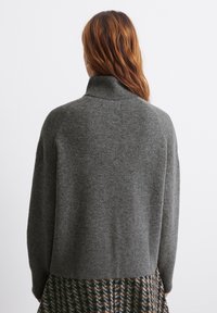 Harmaa neulottu villapaita korkealla kauluksella, jossa on väljä istuvuus ja pitkät hihat, yhdistetty hillityn väriseen kuvioituun hameeseen.