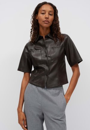 Donna con capelli ricci che indossa una camicia nera in pelle a maniche corte con bottoni e pantaloni grigi a vita alta, in piedi con una mano in tasca.